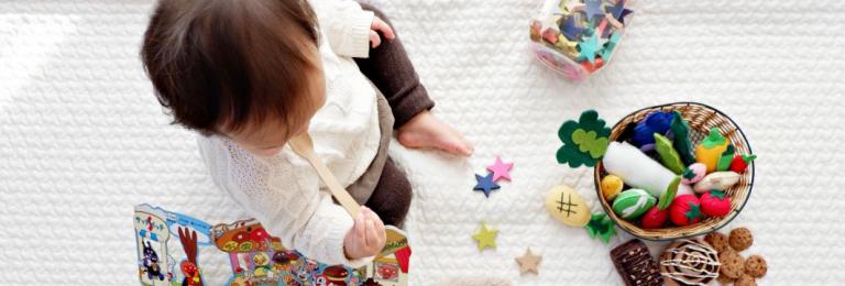 Toddler sitting on white cloth surrounded by toys. Photo by Shitota Yuri on Unsplash.